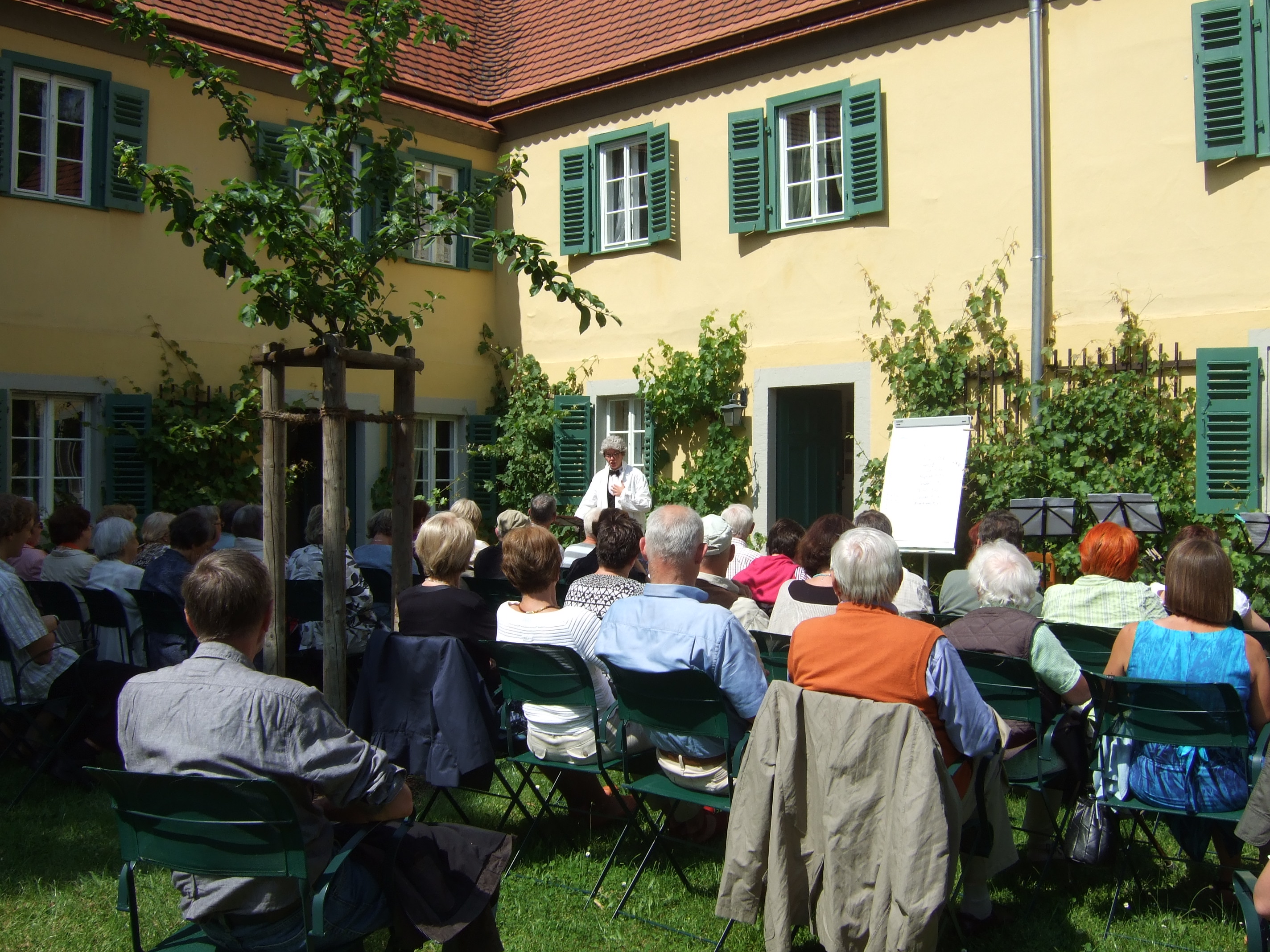 Veranstaltung im Garten des Carl Maria von Weber Museums. Die Gäste sitzen im Garten und schauen zu einer verkleideten Person, die ewtas singt.